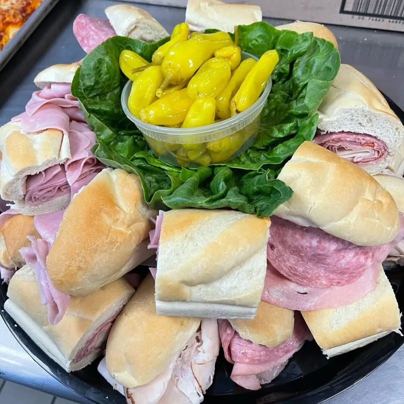 A platter of various deli sandwiches with a side of pickled yellow peppers and lettuce.