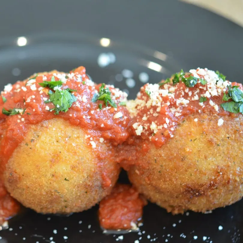 Two golden-brown arancini, topped with red marinara sauce, grated cheese, and fresh parsley.